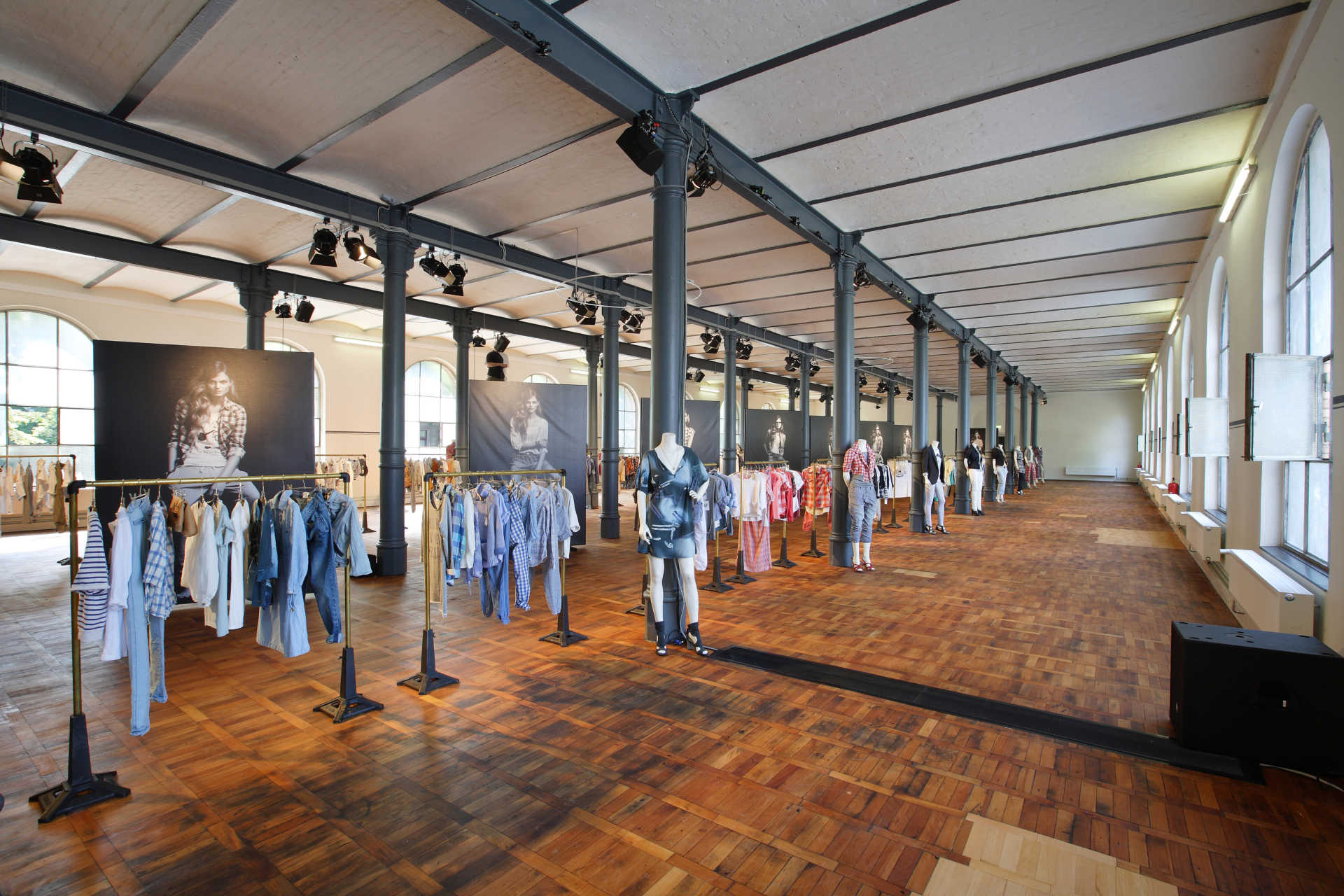 Langer industrieller Saal mit vielen Kleiderstangen, Schaufensterpuppen und großen Postern.