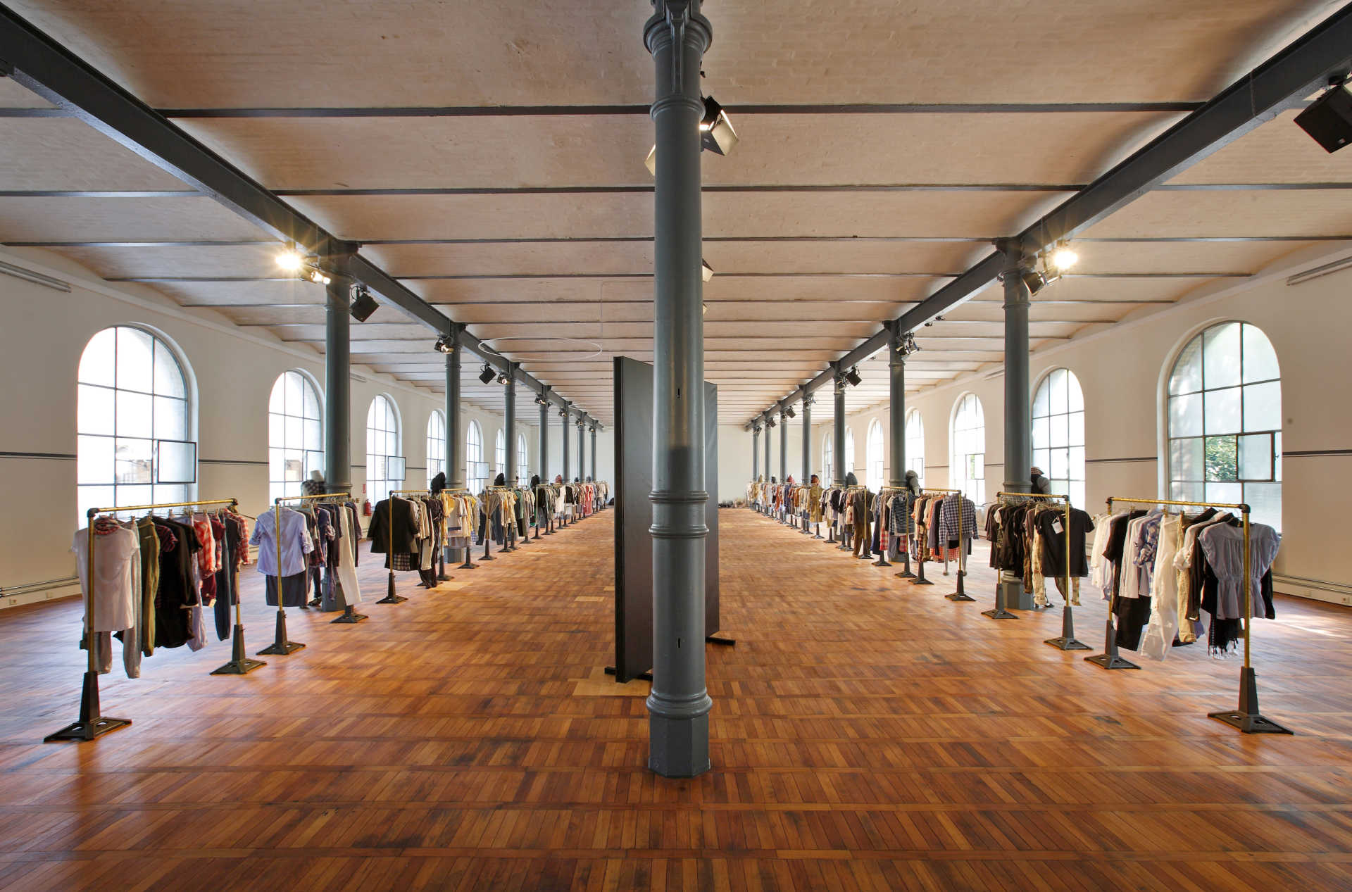Langer industrieller Saal mit Holzparkett, zwei langen Bahnen mit Kleiderstangen und vielen Fenstern.