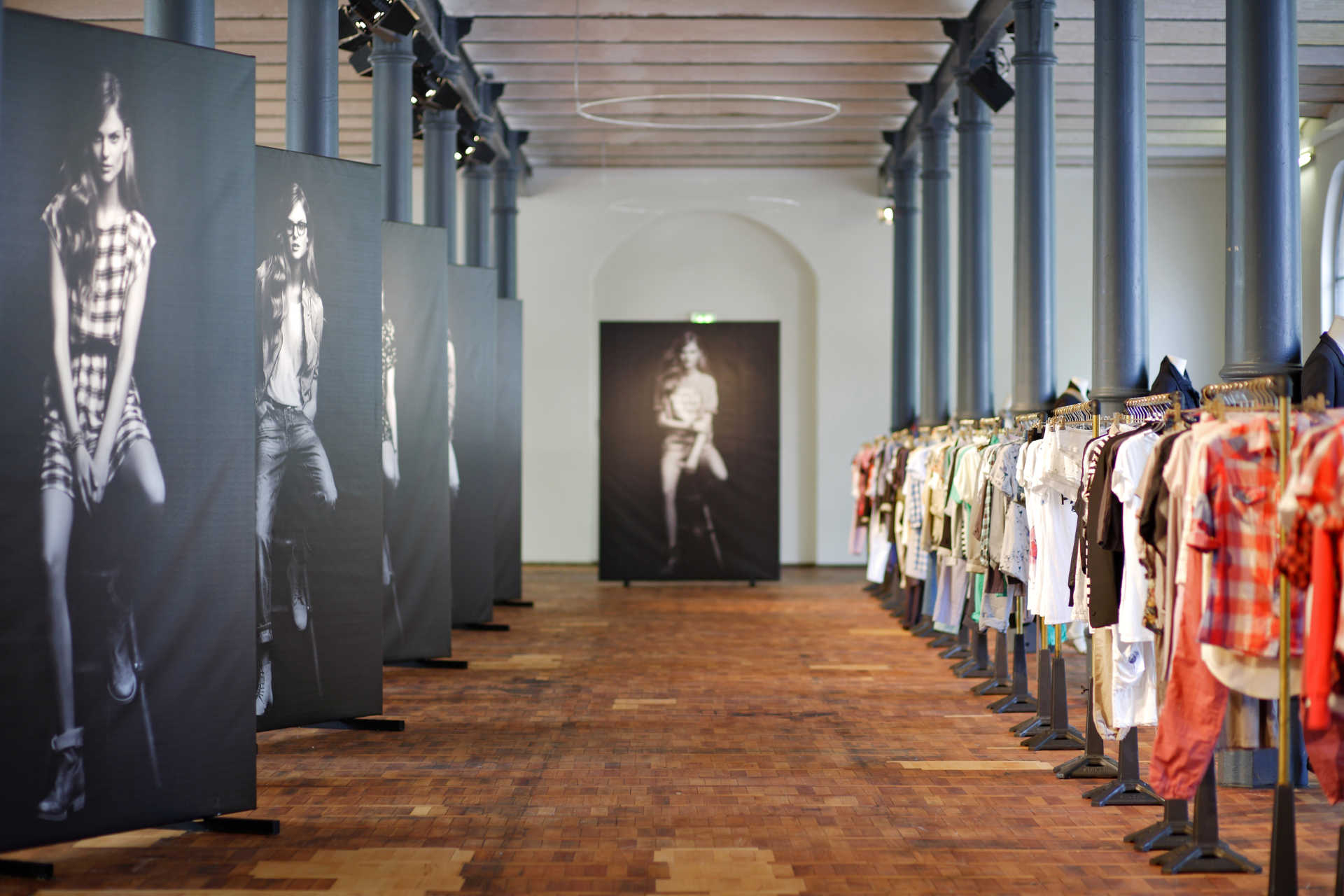Langer Saal mit Holzparkett und langer Bahn aus Kleiderstangen rechts und großen Bannern mit Modefotografien links.