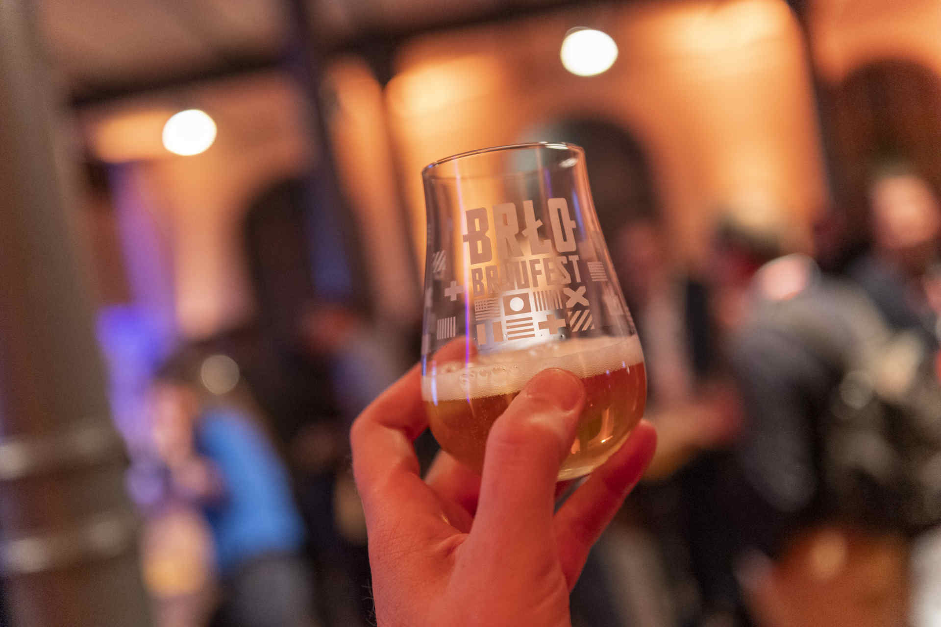 Eine Hand erhebt ein kleines Bierglas im Hintergrund ist unscharf zu erkennen, dass Leute bei einer Abendveranstaltung sind