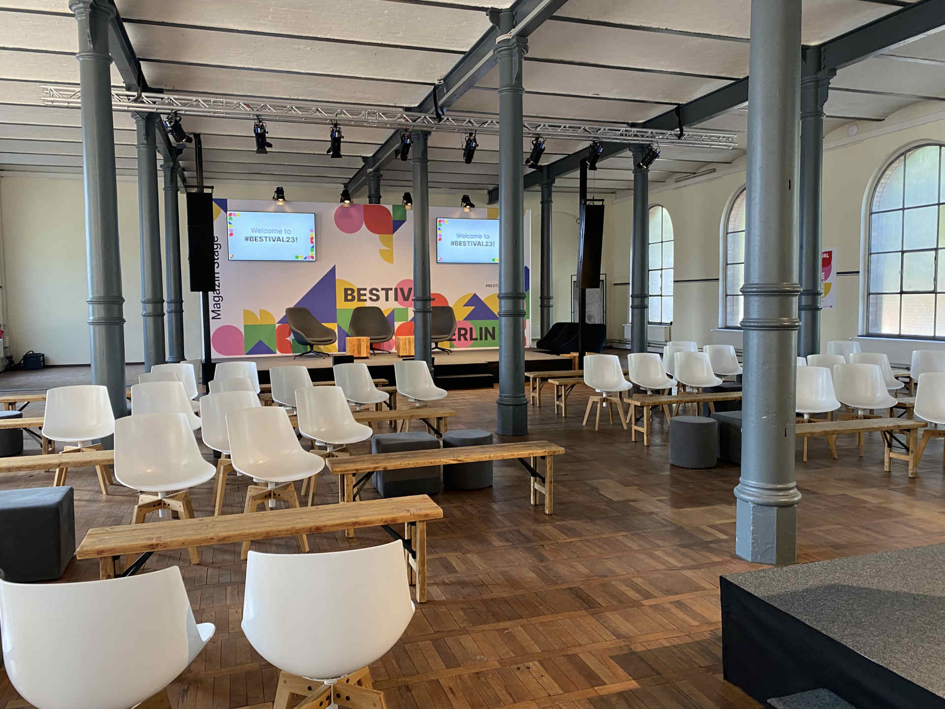 Industrielle Venue mit Säulen, Holzboden, heller Bestuhlung und bunter Pressewand im Hintergrund