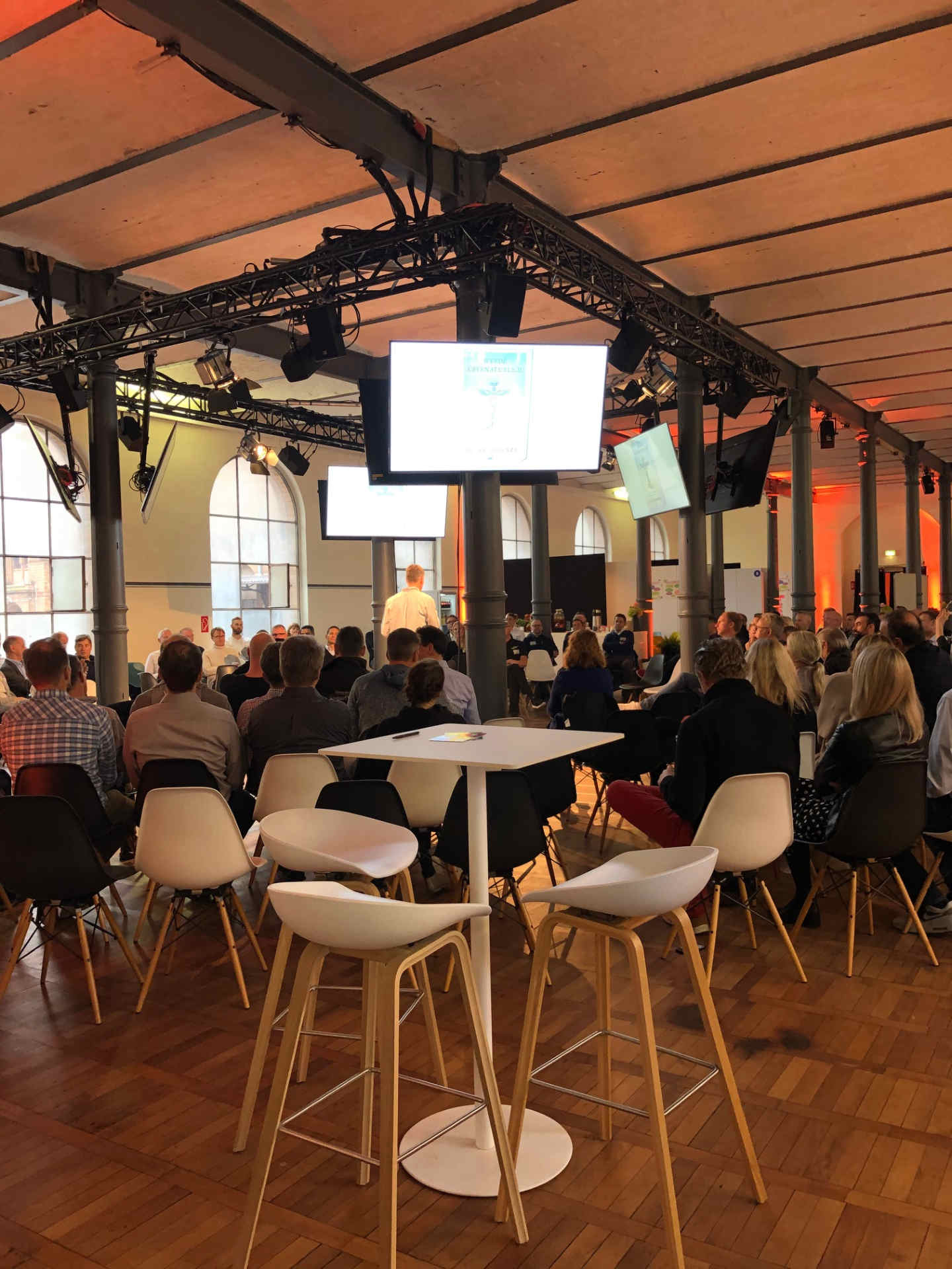 Eventsaal mit vielen Menschen zur Bühne schauend und Monitoren an Säulen