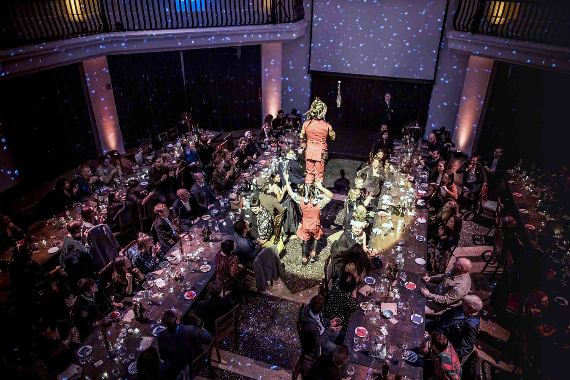 Vollbesetzter Raum mit Menschen an langen Tafeln, Akrobaten in der Mitte